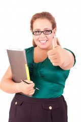 Young lady with red hair, glasses, vibrant green short sleeved top, black jeans smiling and holding a clipboard while giving a thumbs up!